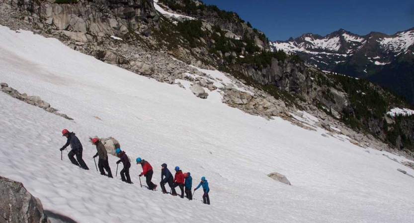 A group of people wearing mountaineering gear hike in a line up a snowy incline